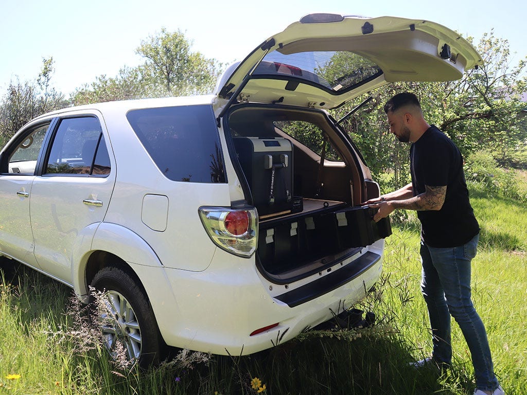 A man opens the back of a white Toyota Fortuner fitted with a Front Runner Wolf Pack Pro storage system, in green bush