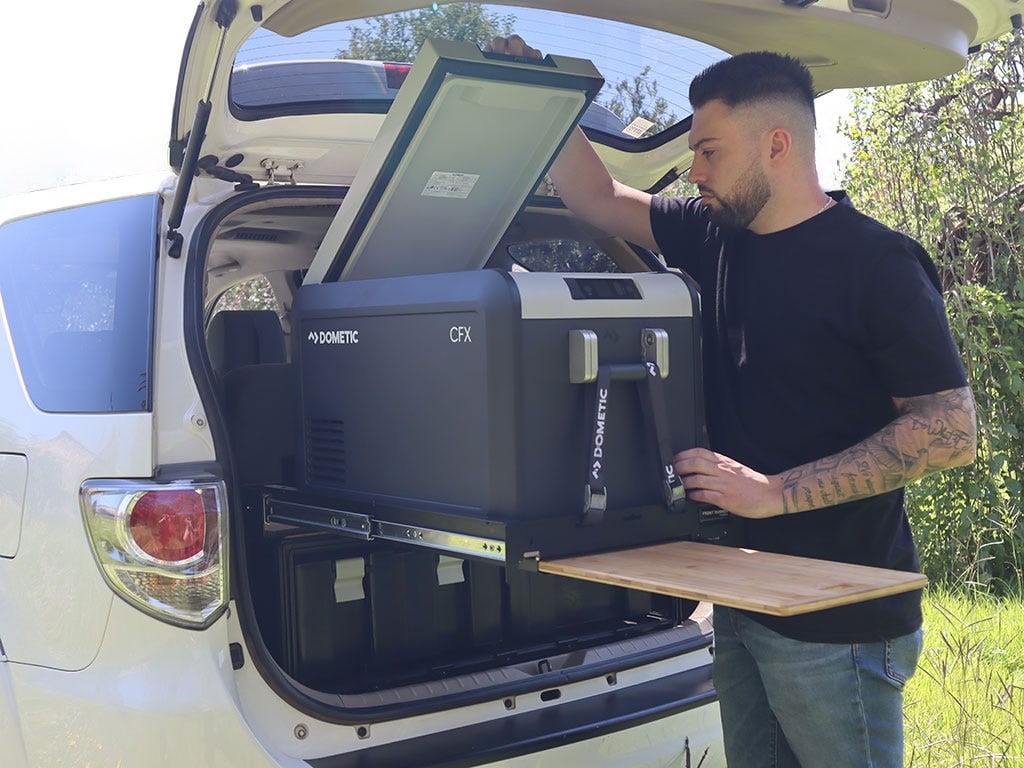 A man opens a Dometic fridge on a pull-out slide on top of a Wolf Pack Pro storage system in the boot of a white Fortuner
