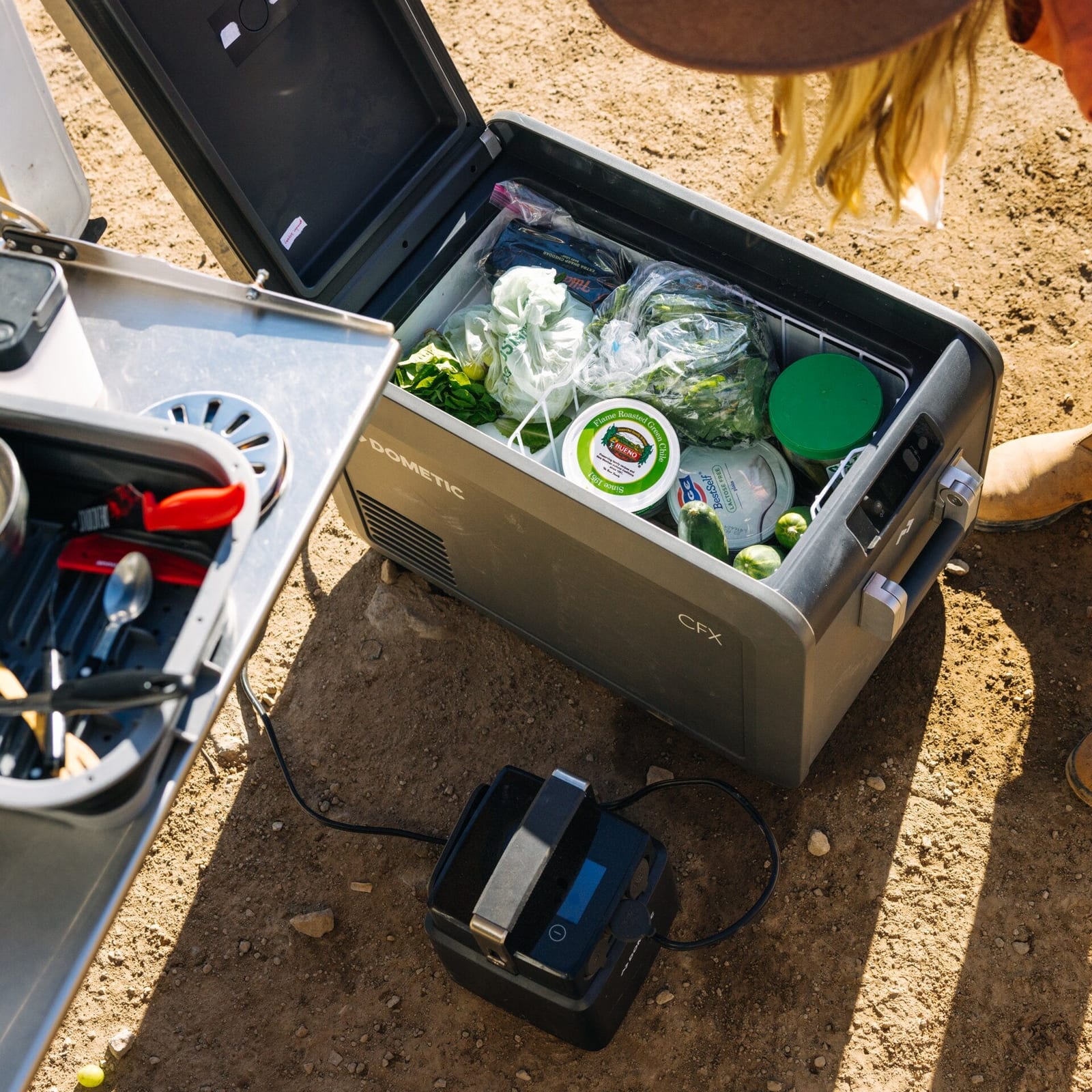 A Dometic CFX5 fridge full of food, lid open, on dusty ground next to a camp kitchen and a portable battery