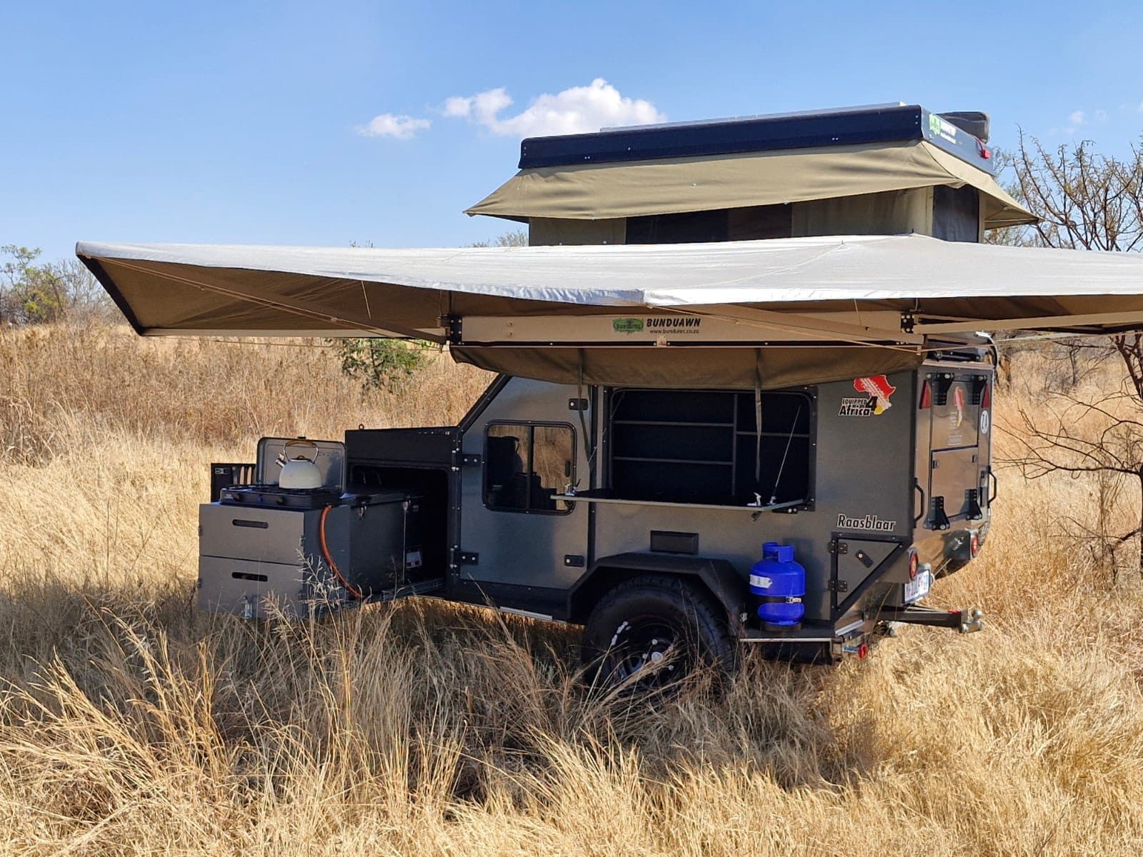 Fully-equipped off-road camper trailer set up with rooftop tent, awning and camp kitchen in the South African bush