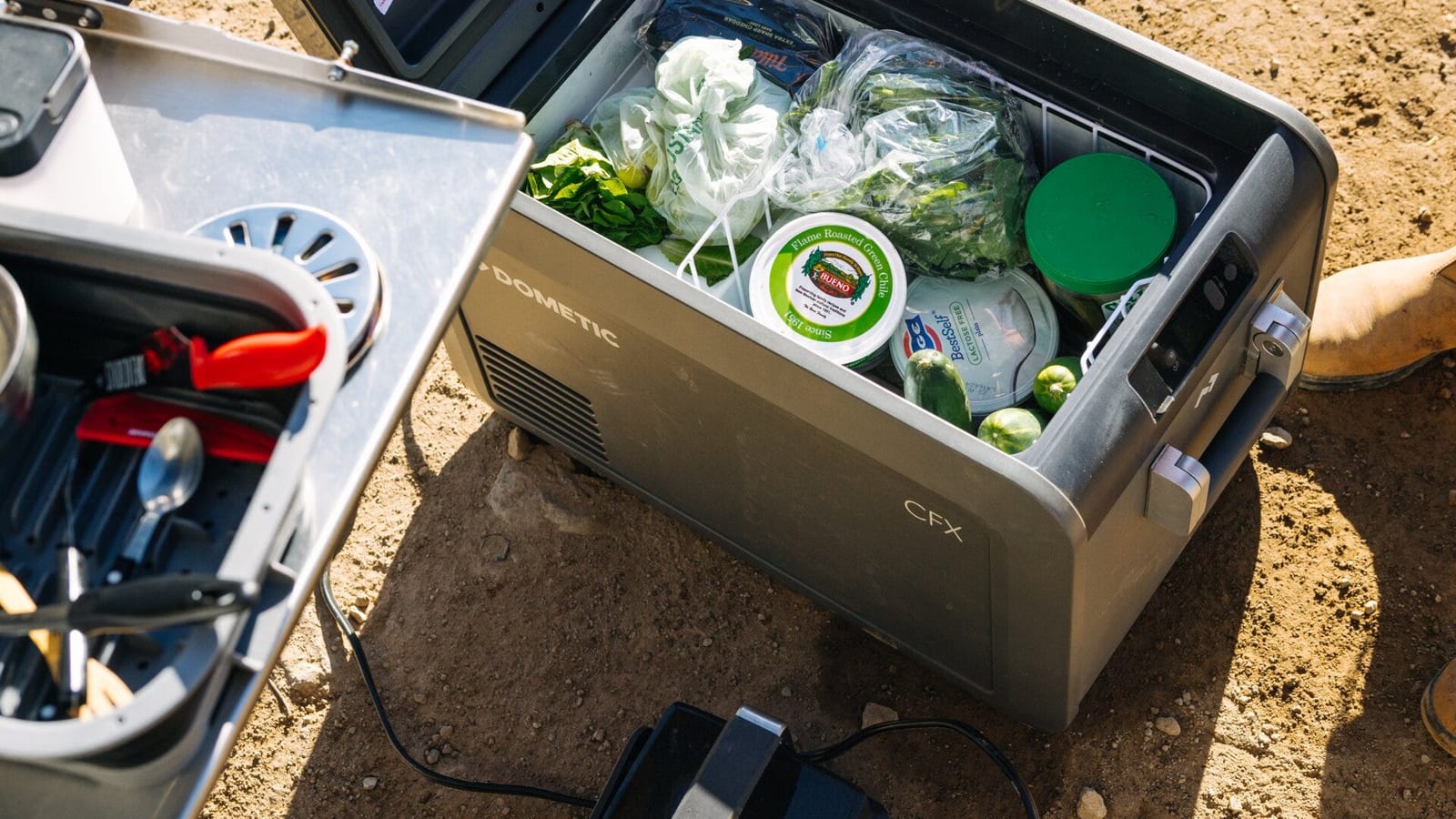 A Dometic CFX5 fridge full of food, lid open, on dusty ground beside a camp kitchen