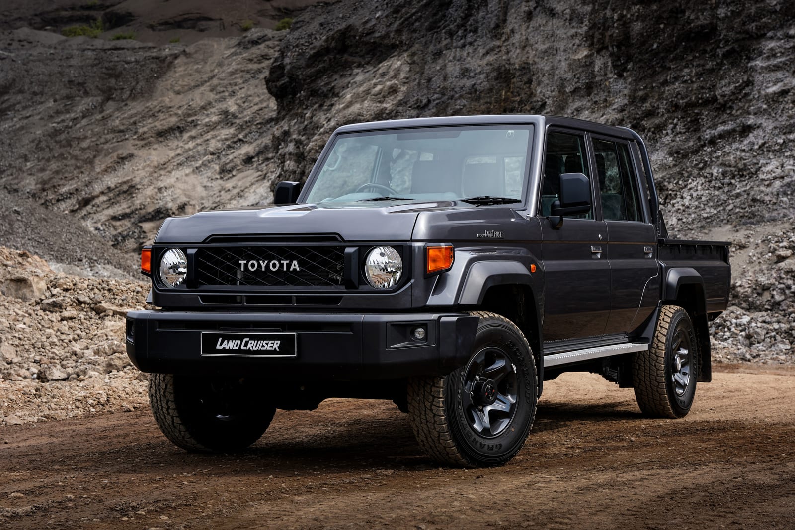 A dark grey Toyota Land Cruiser 79 double cab parked on a dirt track in front of a rock face.