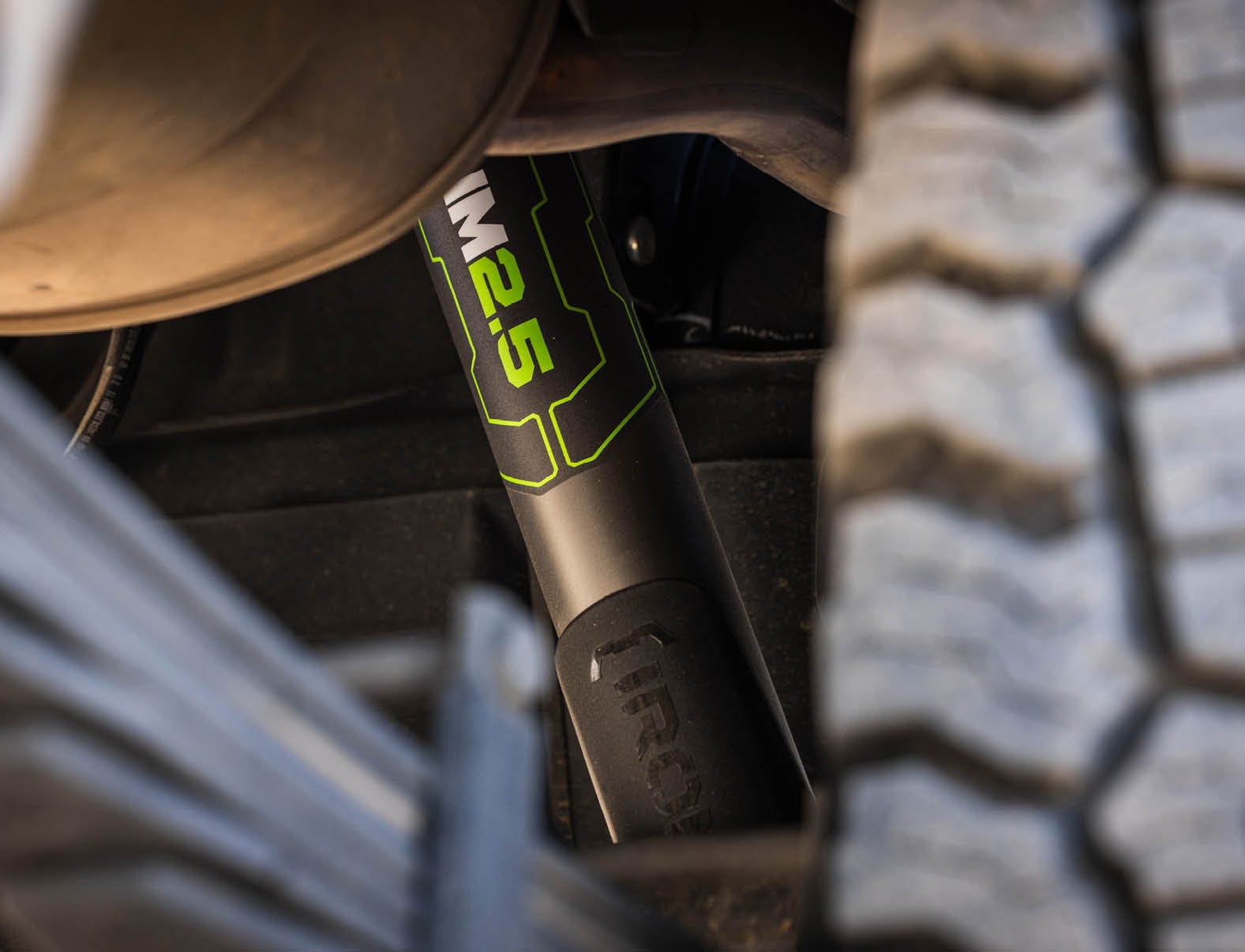 A close-up of a black and green monotube shock fitted next to a rear leaf spring pack and an off-road tyre