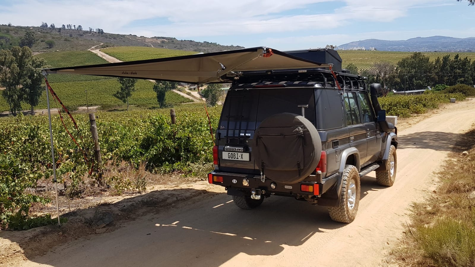A kitted Land Cruiser 76 station wagon parked on a farm track between vineyards with its side awning rolled out