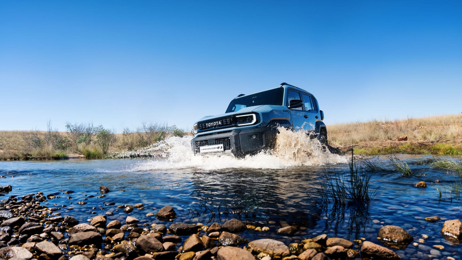 A new Toyota Land Cruiser FJ splashing through a shallow, rocky river on dry veld.
