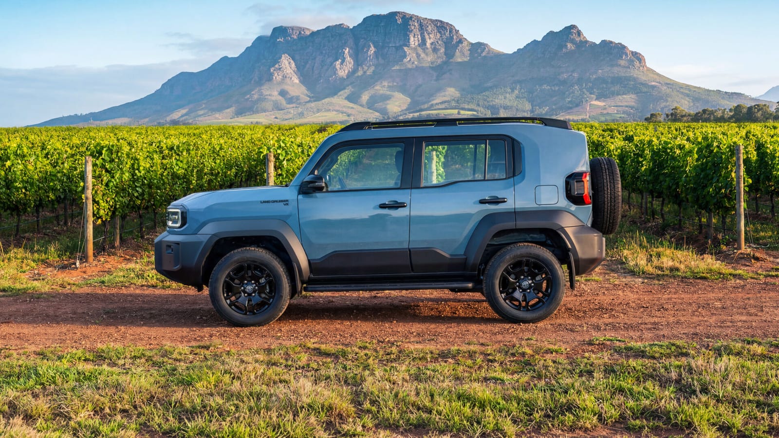 Side view of a new Land Cruiser FJ on a dirt track between vineyards, mountain behind.