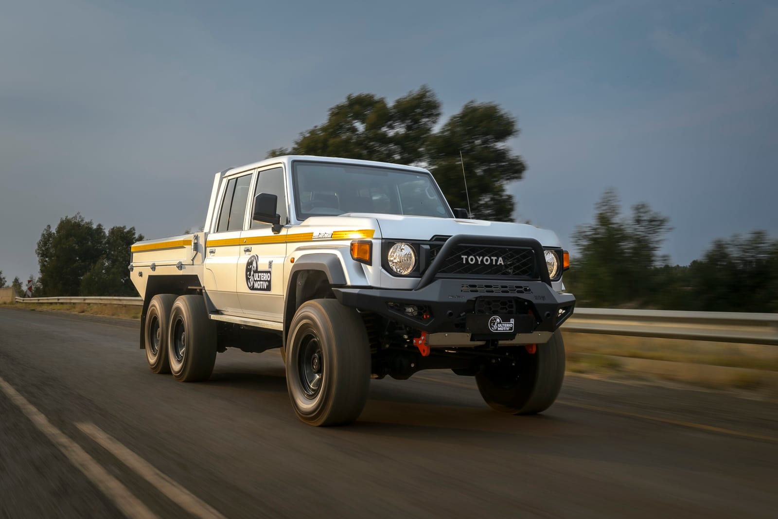 A white Land Cruiser 79 double cab with six wheels and a flat load bed driving on a South African road