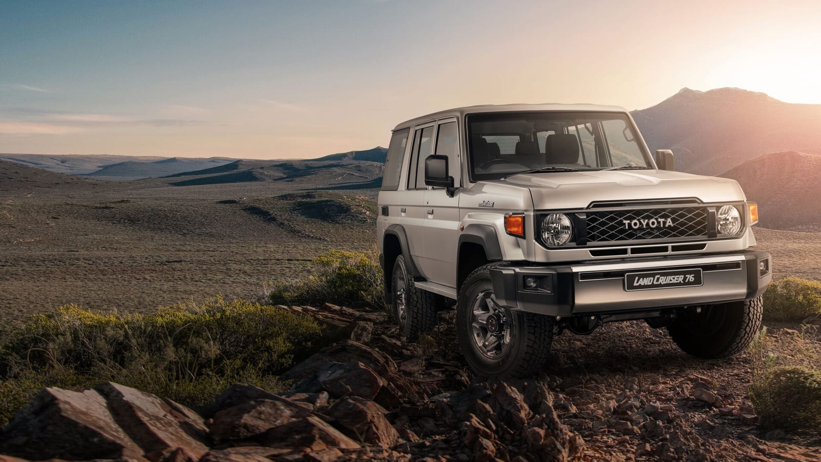 A new-shape Toyota Land Cruiser 76 station wagon parked on a rocky rise above wide empty valleys at sunset