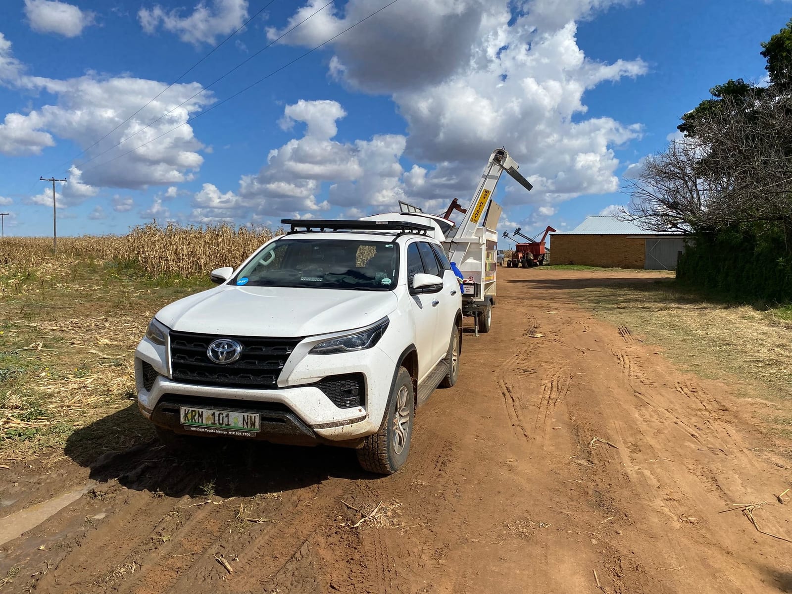 A white Toyota Fortuner towing a farm trailer on a dirt road next to a mielie field.