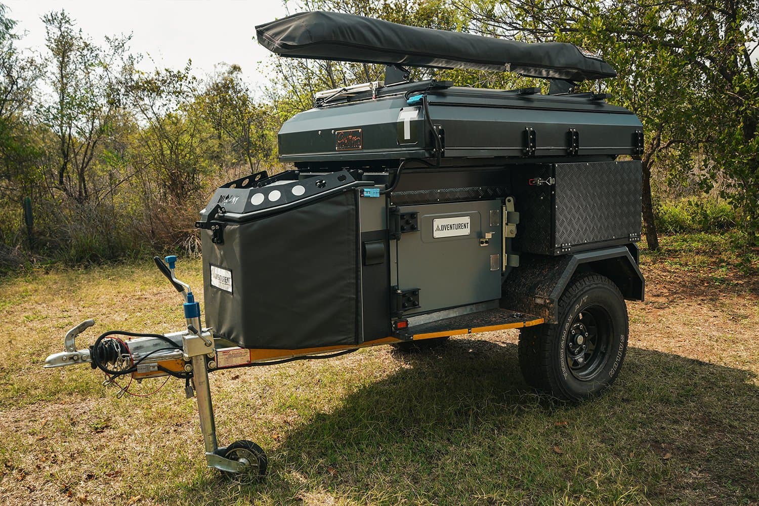 A sleek off-road camper trailer with a closed rooftop tent parked in the bushveld