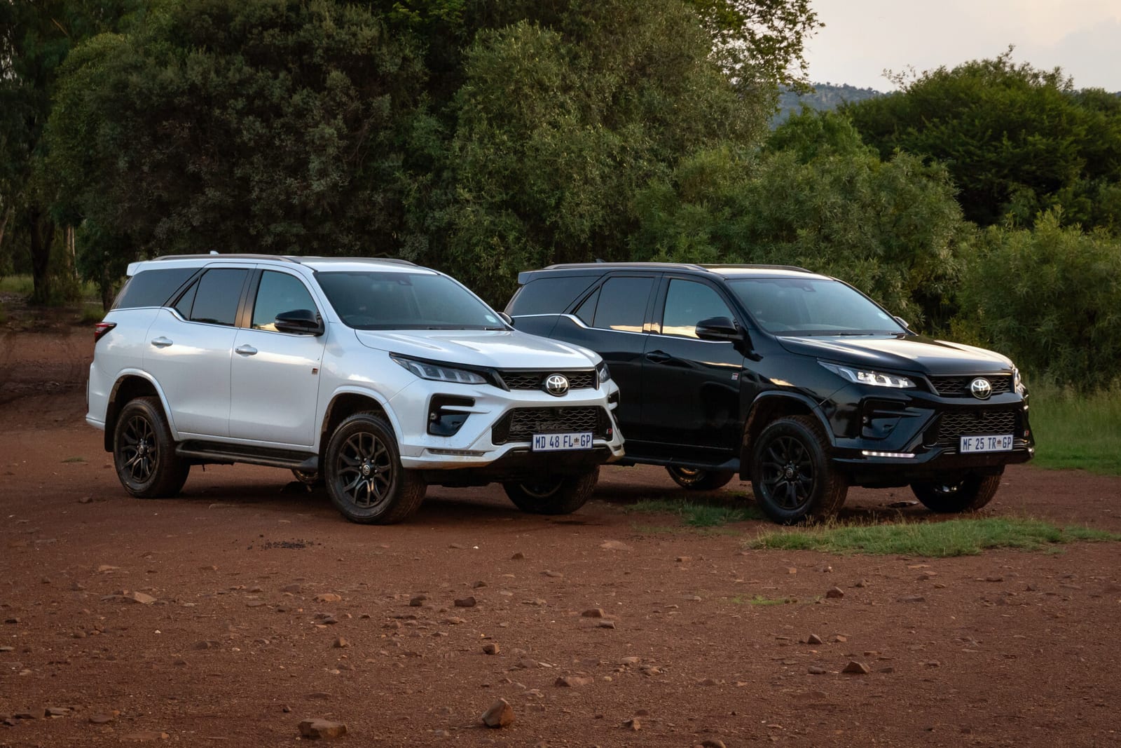 A white and a black Toyota Fortuner GR-S parked on a red dirt clearing under trees.