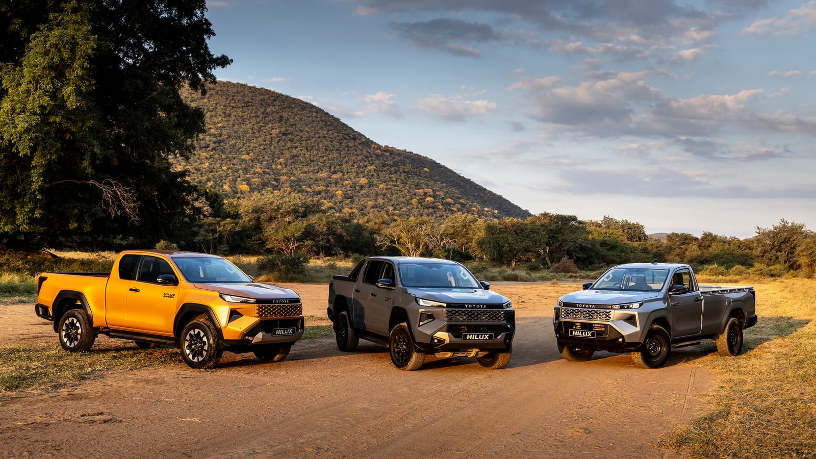 The new 2026 Toyota Hilux in orange Xtra Cab, grey Double Cab and silver Single Cab versions parked on a dirt road in South Africa.