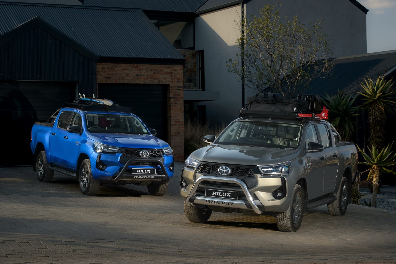 A blue and a grey Toyota Hilux Raider double cab with nudge bars and roof racks parked in a driveway.