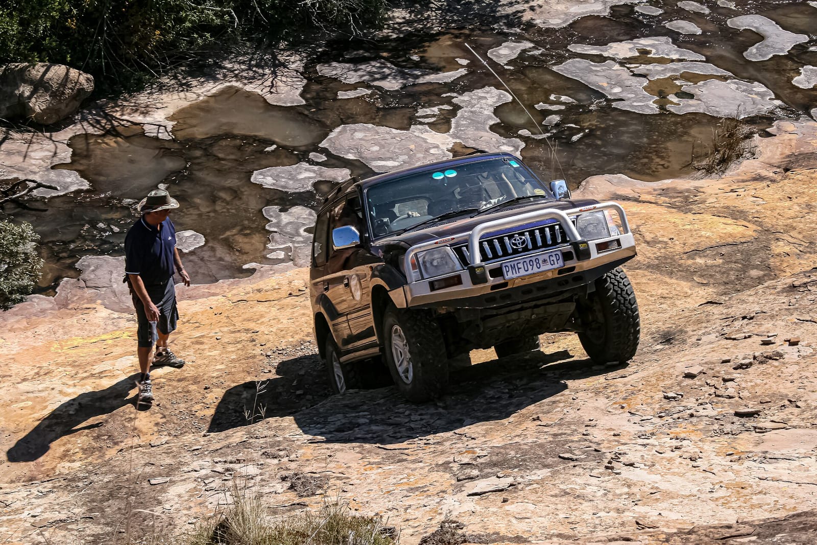 A dark Toyota Prado 90 with a bull bar climbing over sandstone, with a man walking beside it.